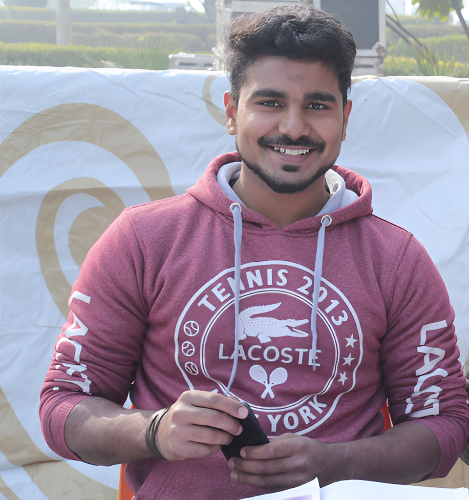 The image shows a young man smiling warmly at the camera. He has dark hair and a well-groomed beard, and he is wearing a maroon hoodie with white drawstrings and the text "TENNIS 2013 LACOSTE NEW YORK" printed on the front, along with a graphic of a crocodile and tennis-related symbols. He is holding a black smartphone in his hands, and the background appears to be an outdoor setting with some greenery and a white banner featuring circular patterns behind him.
