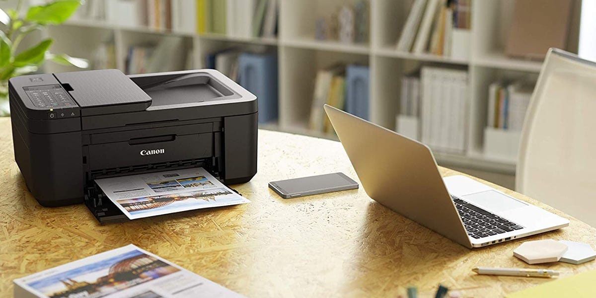 A Canon printer on a desk with a laptop beside it, a print coming out. 