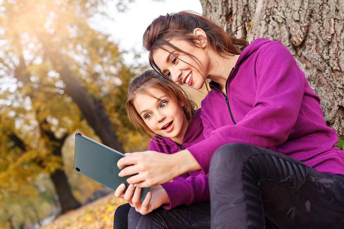 Woman and girl using tablet while sitting against a tree