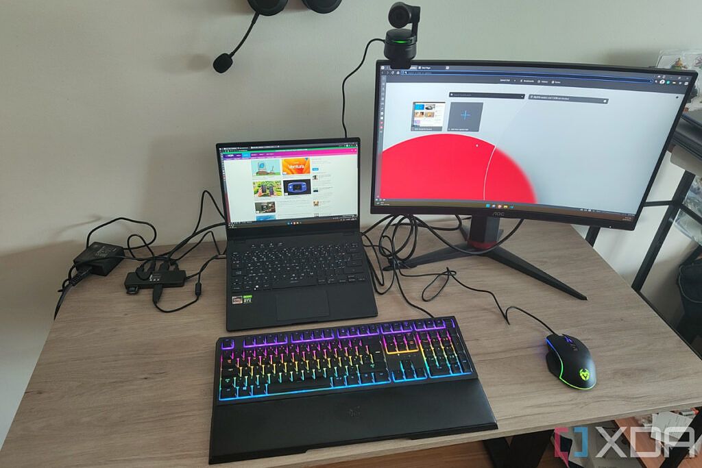 Un escritorio configurado con una computadora portátil, un monitor, un teclado, un mouse y una cámara web USB, todos conectados a un concentrador, que también carga la computadora portátil.