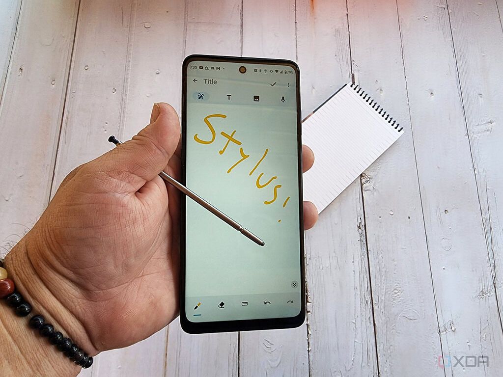 A man's hand hold the Moto G Stylus 5G over a table with a note pad on it.