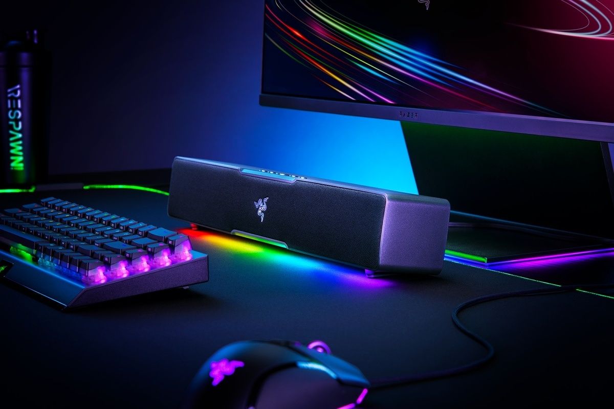 Angled view of the Razer Leviathan V2 X soundbar with RGB lighting enabled, next to other peripherals includinga keyboard, mouse, and monitor