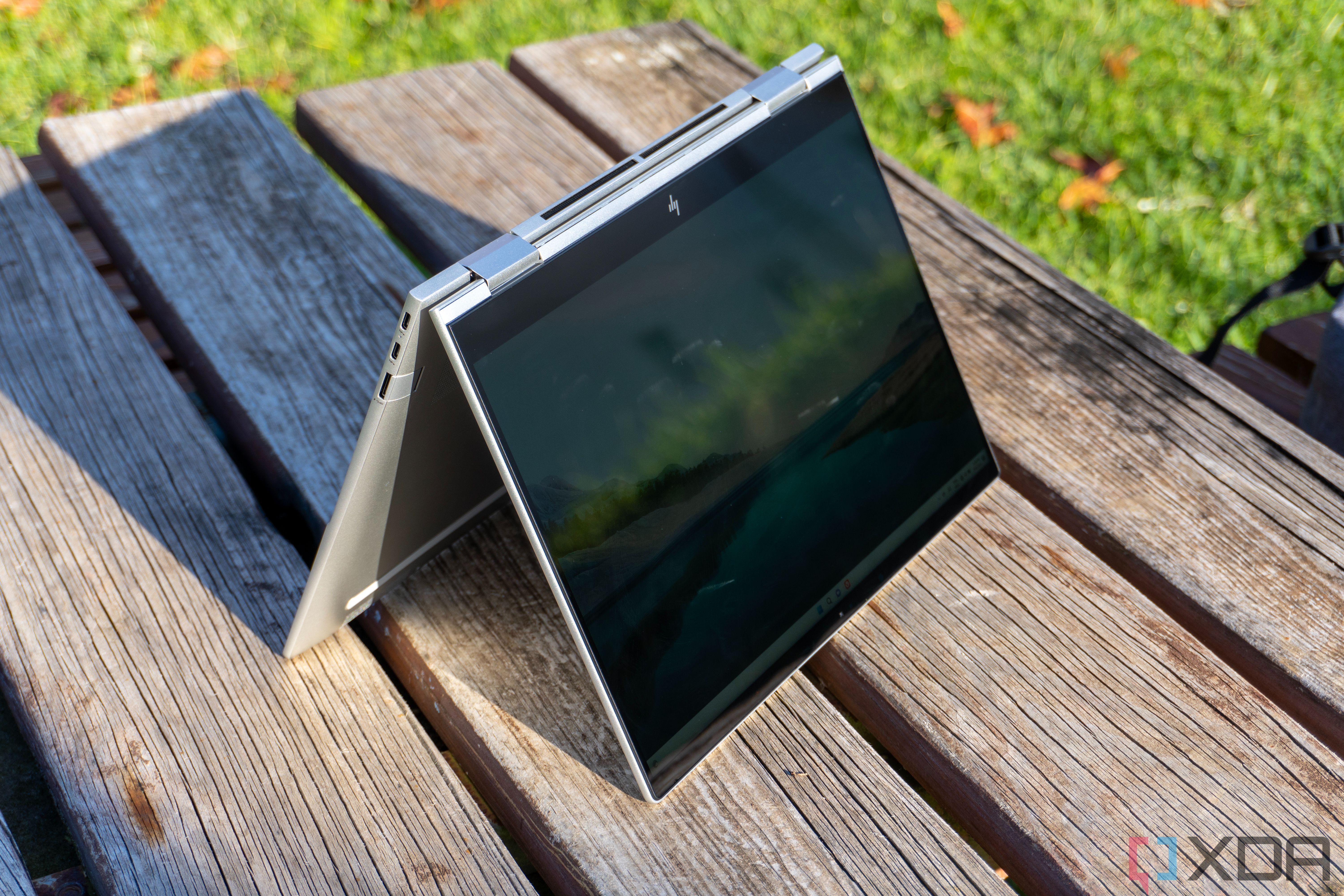 HP Envy x360 13 in tent mode, seen at a left-side angle with the screen directly facing the sun. THe laptop is standing on a wooden table.