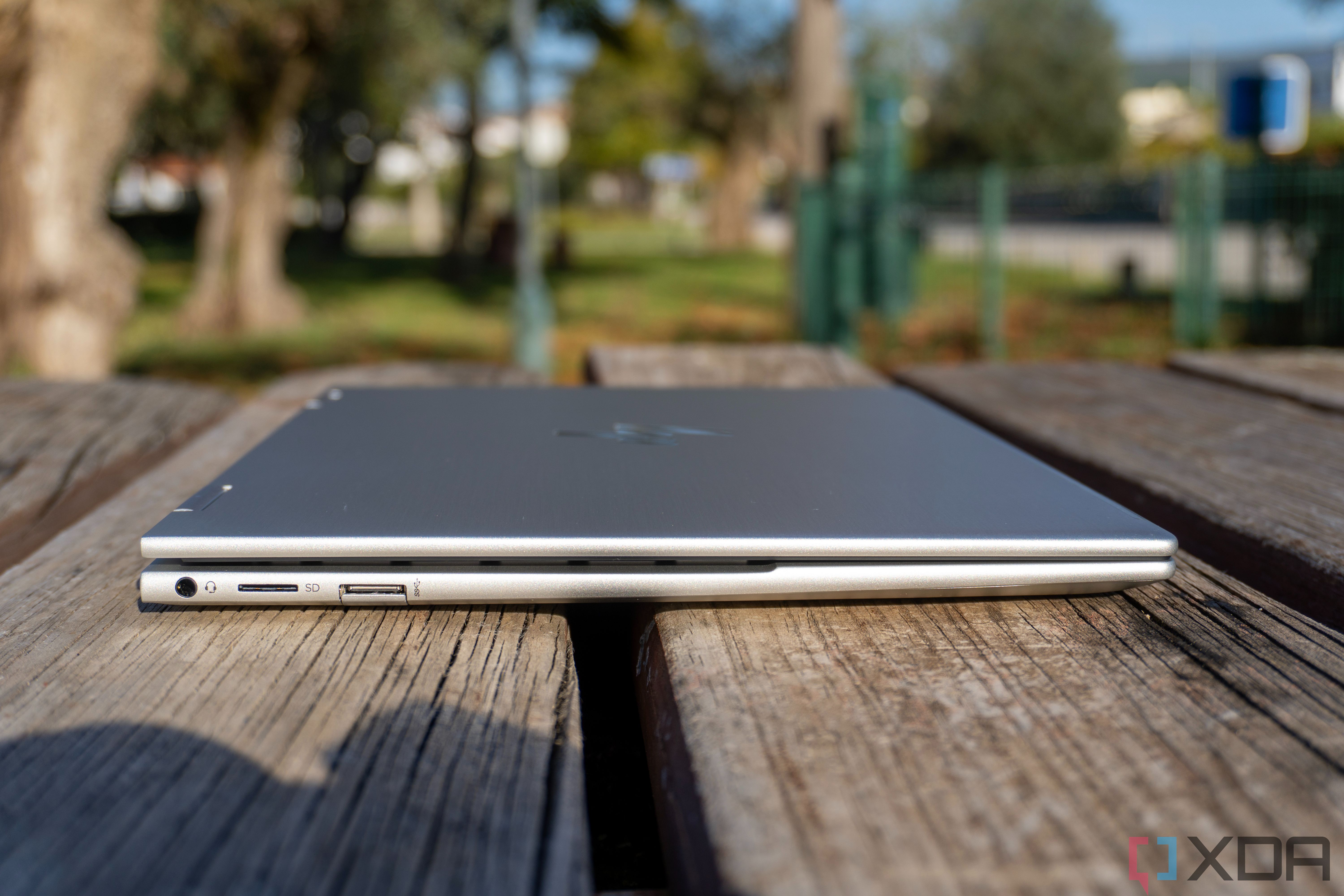 Left-side view of the HP Envy X360 with the lid closed, showing off a headphone jack, microSD card reader, and a USB Type-A port. The laptop is placced on wooden boards and you can see a park in the background