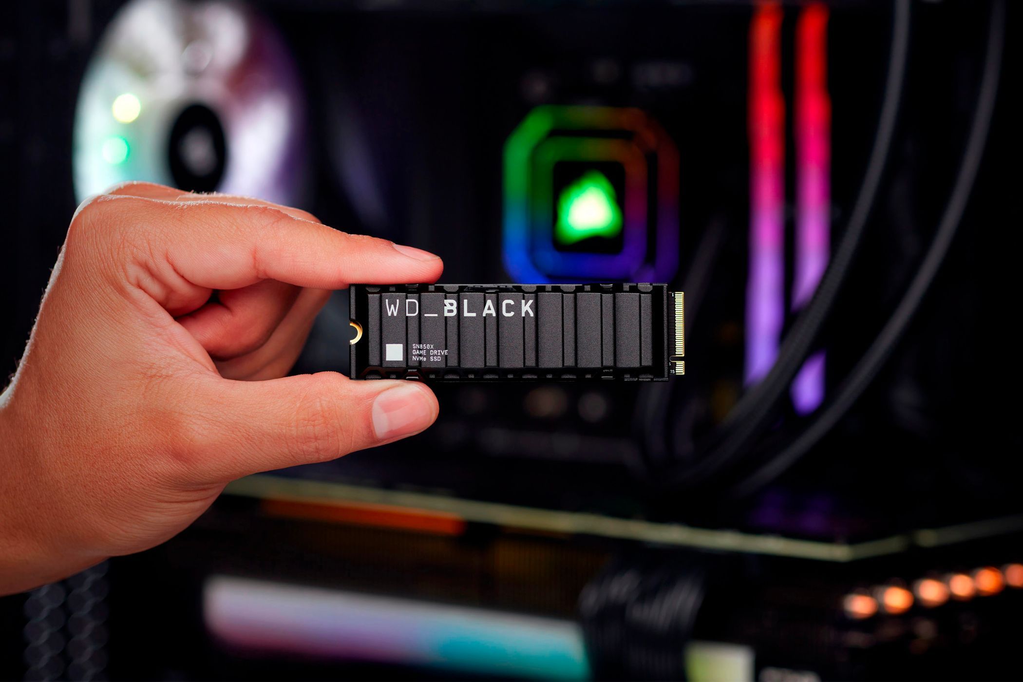 A person holding the WD Black SN850X gaming ssd with their thumb and the index finger with a gaming PC with RGB lights in the background.