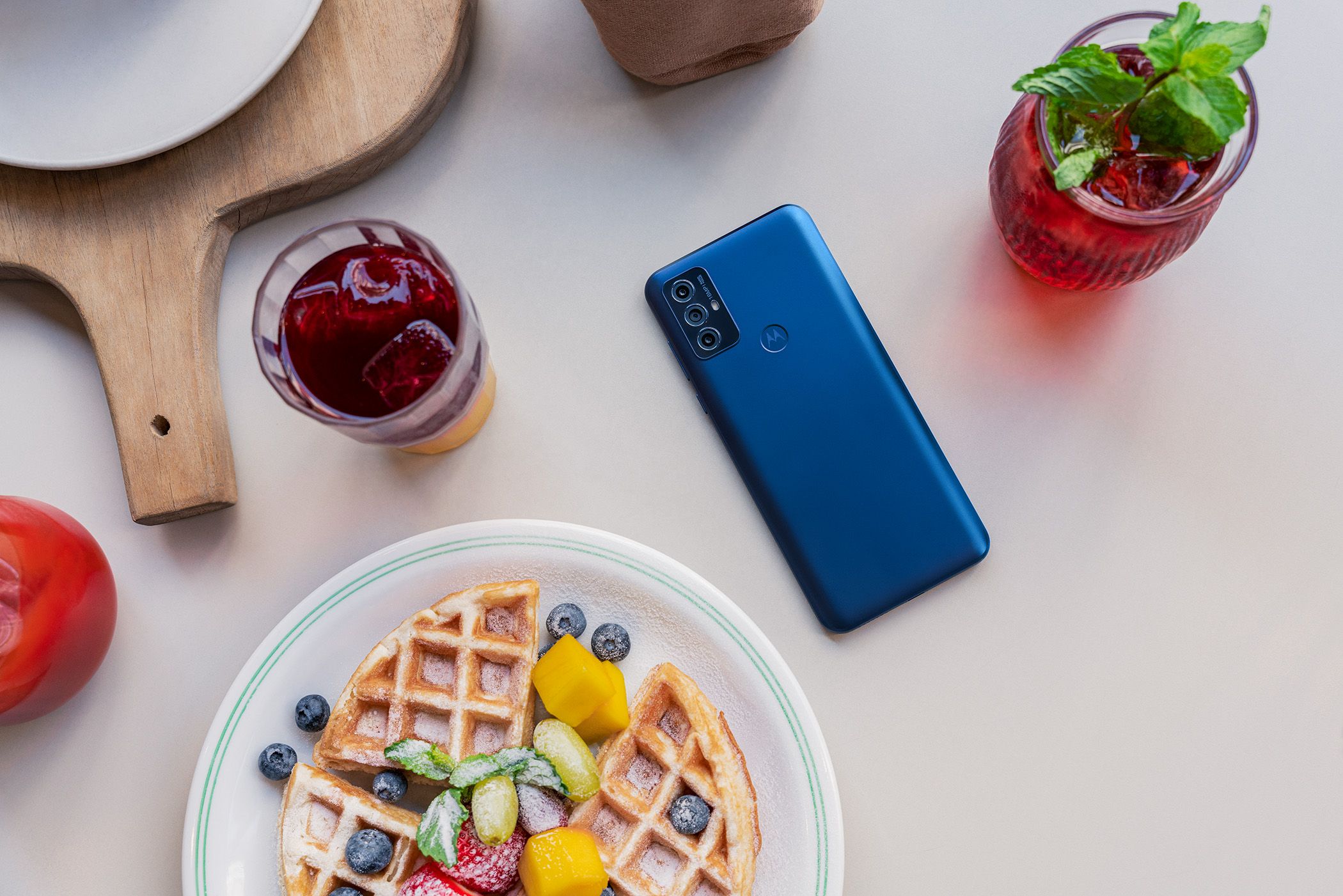 The Moto G Play 2023 is placed face down on the table next to drinks and pies.