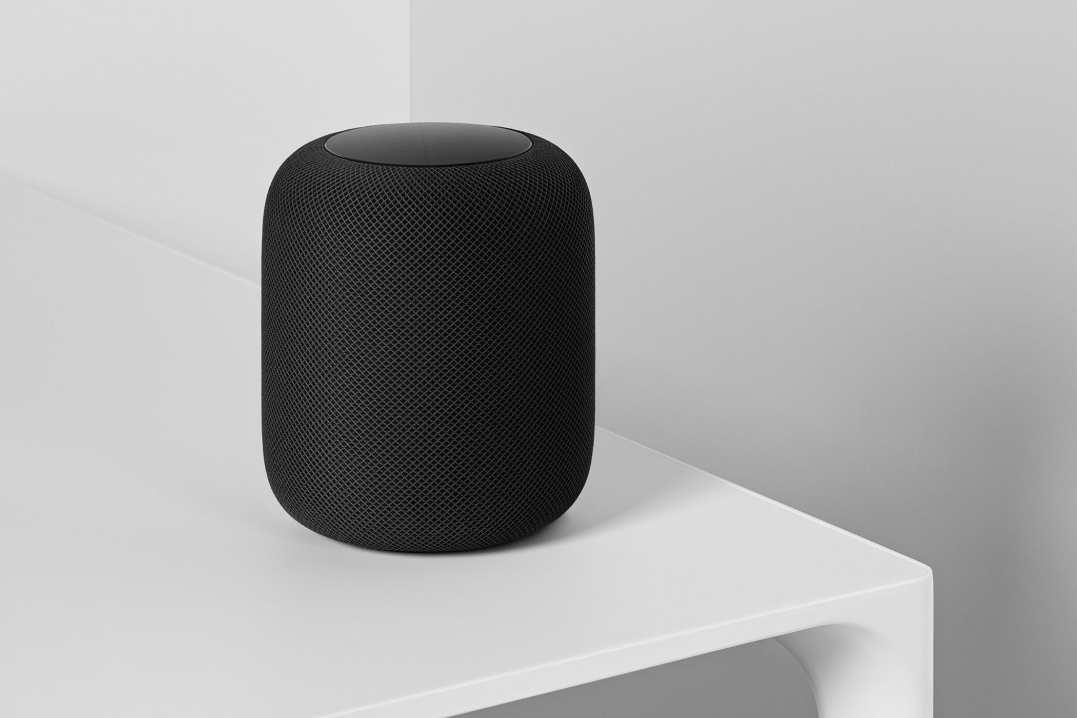 Apple HomePod negro sobre una mesa blanca con paredes blancas de fondo.