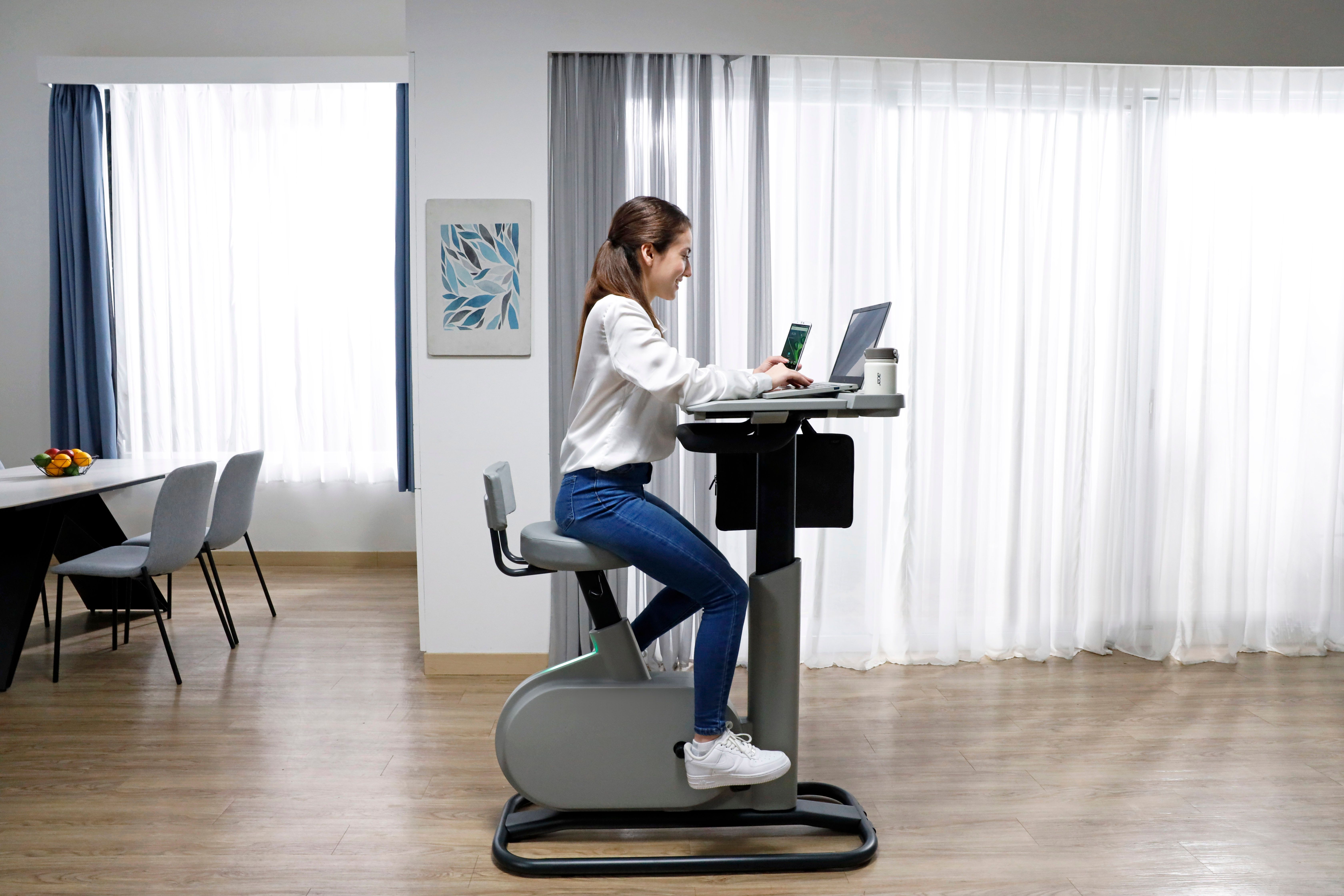 A woman sitting on the eKinect Bike Desk with the desk pulled closer to the seat. The desk has a laptop and a phone on it, which the woman is using to work.