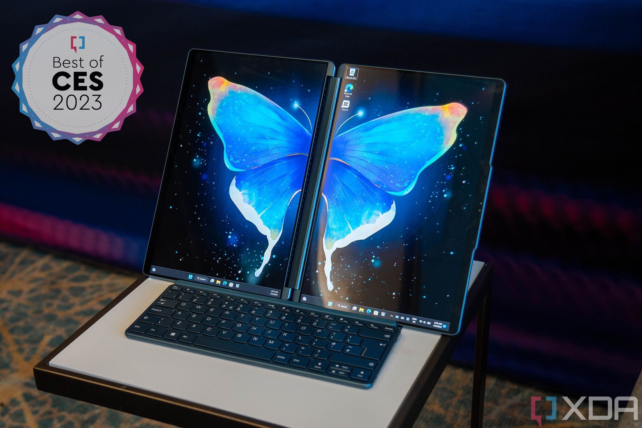 A Lenovo Yoga Book 9i laptop with two screens in portrait orientation, which line up with each other to make a larger landscape display. A keyboard is attached at the bottom. In the top left corner, there's a badge with text reading "Best of CES 2023"