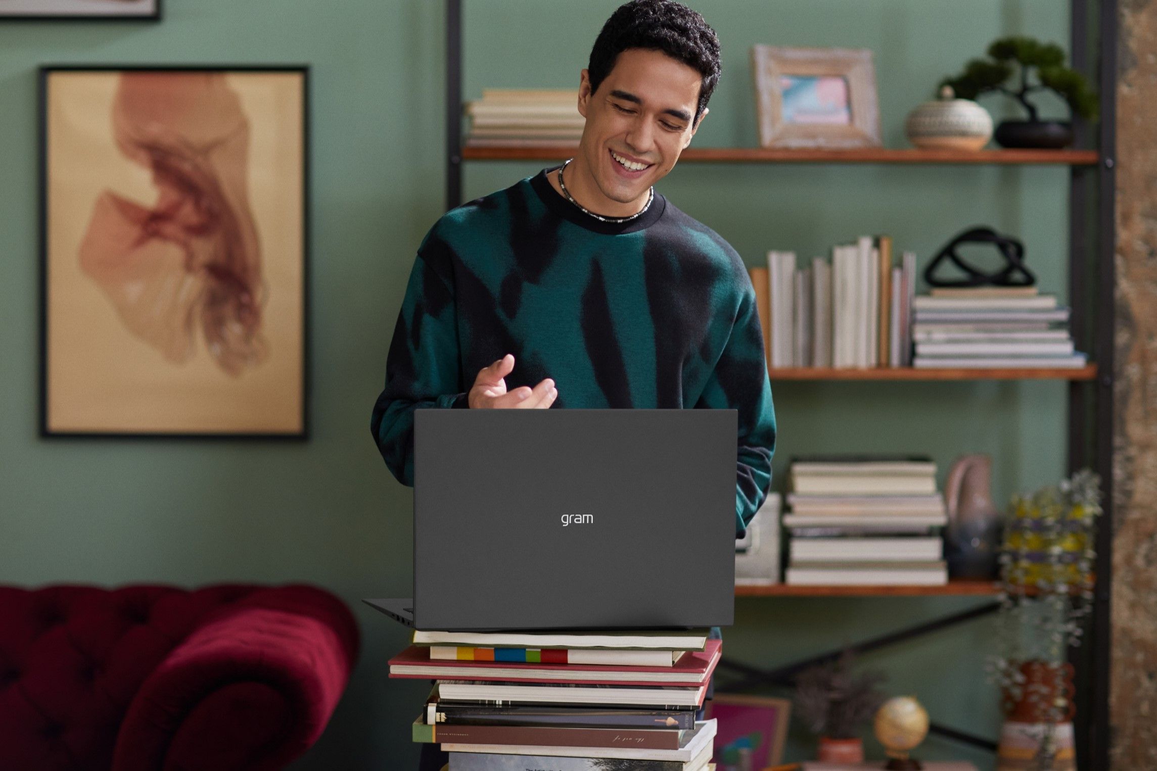 An LG gram laptop sitting on a pile of books, facing away from the camera. A man is standing in front of the laptop and smiling.