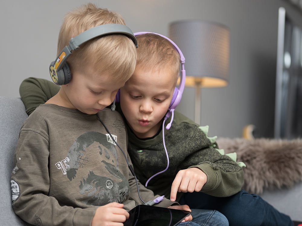 Two childdren wearing Onanoff BuddyPhones Cosmos+ headphones with one pair daisy-chained to the other so they can listen to the same tablet