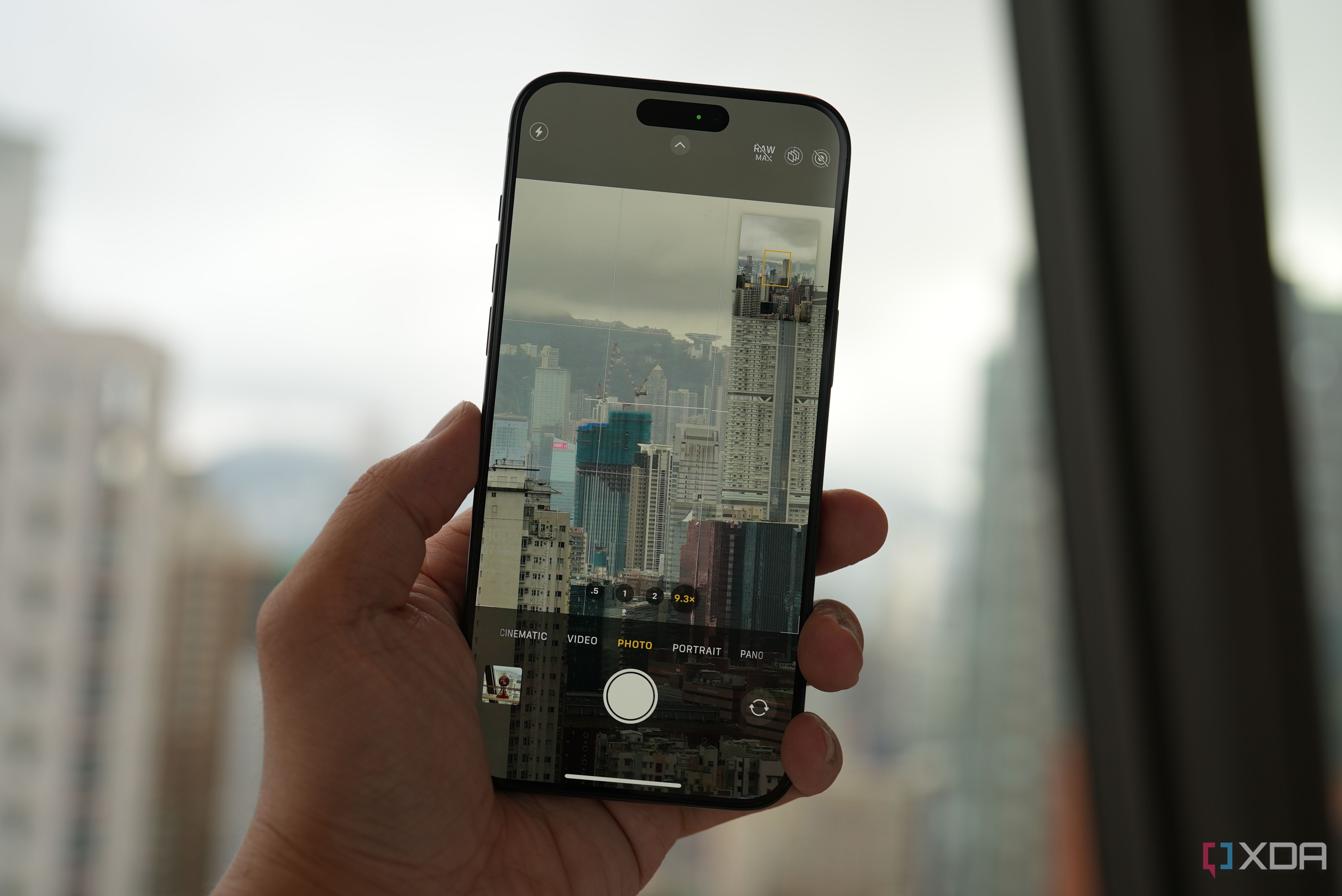 An iPhone 15 Pro being held in a hand taking a photo of a dense city full of high-rises on a cloudy day through a window.
