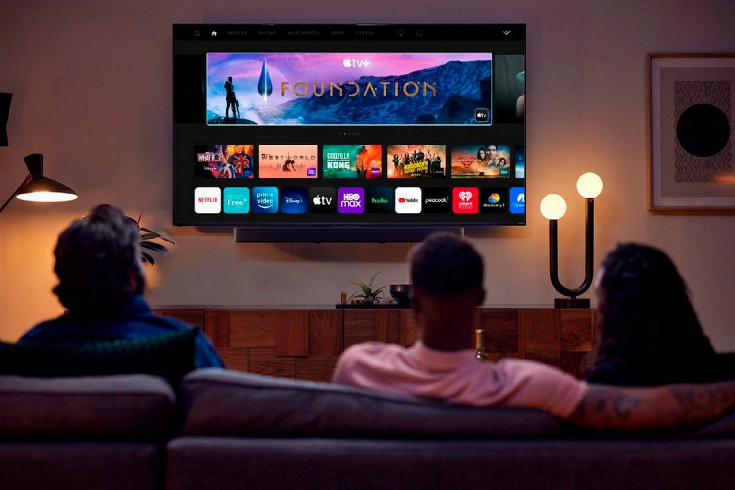 Three people on a couch, pictired from the back, watching a TV mounted on the wall