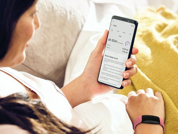 A woman lying bed wearing a Fitbit on her right wrist and looking at her sleep statistics on her smartpgone held in her left hand.