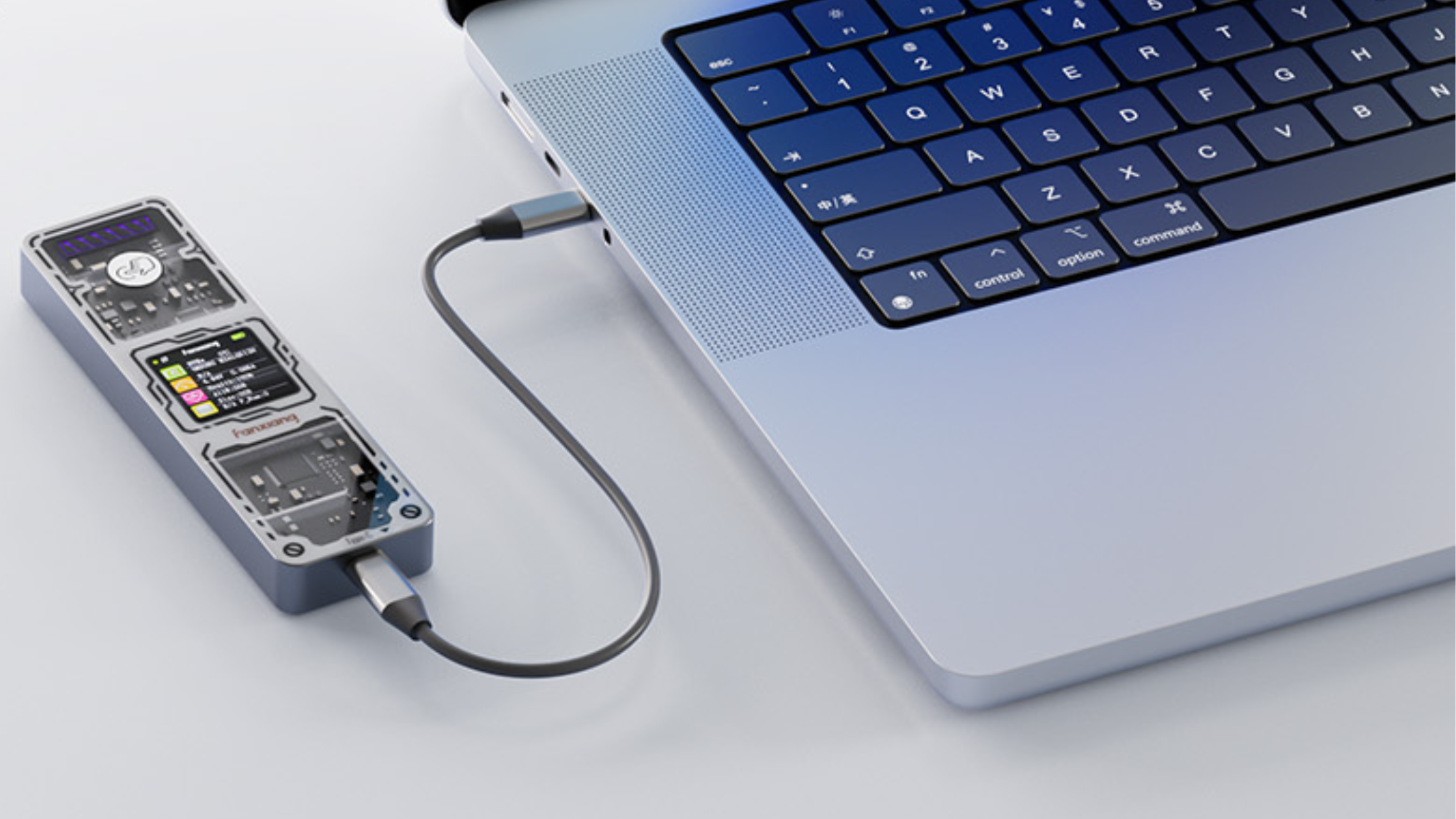 The image shows a close-up view of a modern laptop with a metallic silver body and a black keyboard featuring a compact layout, including keys such as control, option, and command. Connected to the laptop via a short black USB-C cable is a transparent electronic device that reveals internal circuitry, a small screen displaying colored icons and text, and some mechanical components. The device is resting on a light-colored surface, and the overall setting appears clean and minimalistic.