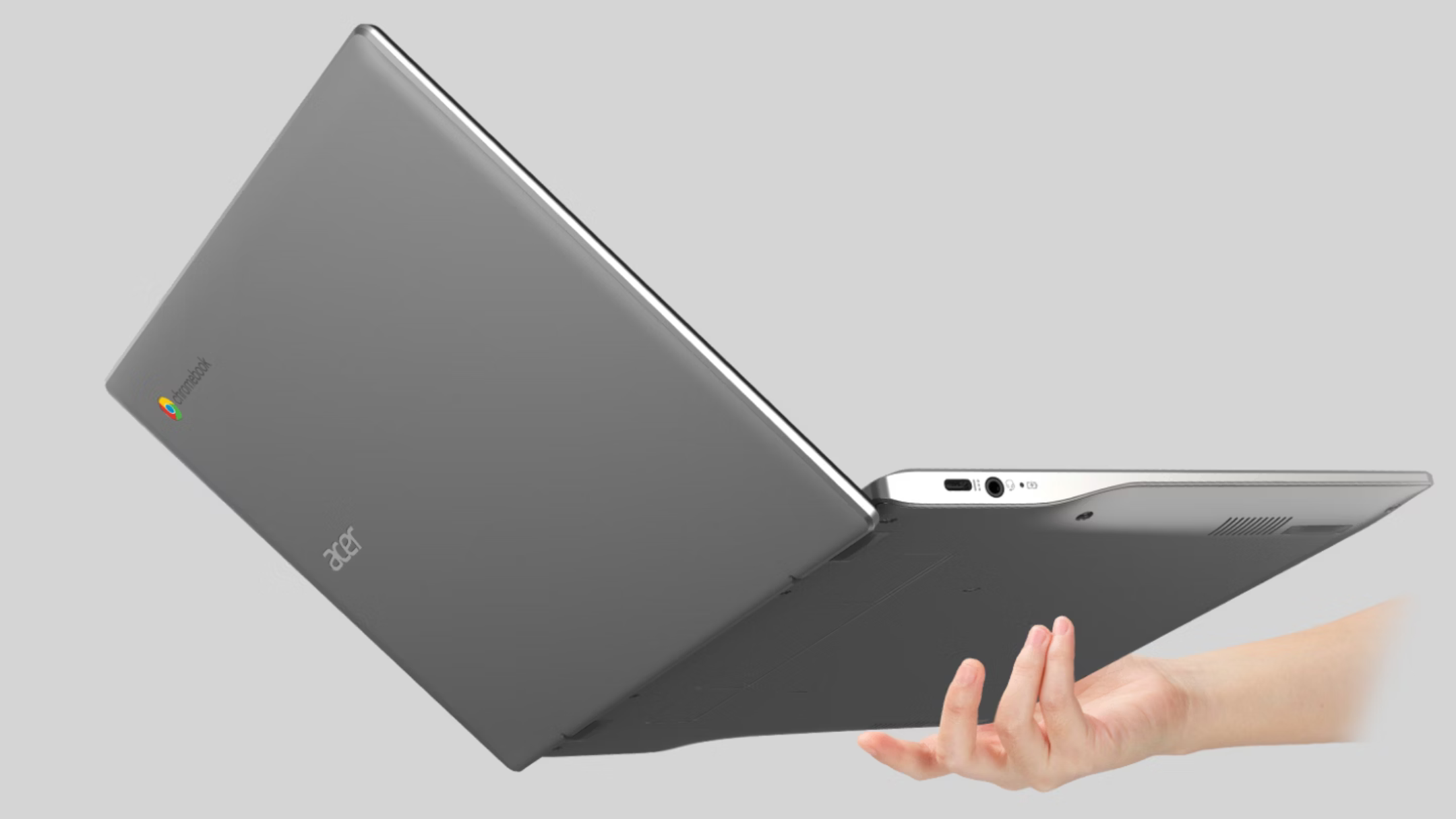 The image shows a sleek, silver Acer Chromebook laptop being held mid-air by a single hand from underneath. The laptop is slightly open, displaying the underside and exterior back of the screen. The Acer logo and a small, colorful Chrome logo are visible on the laptop’s lid, while ports including a USB-C and headphone jack are visible on the side. The background is plain and light gray, drawing full focus to the laptop and hand.