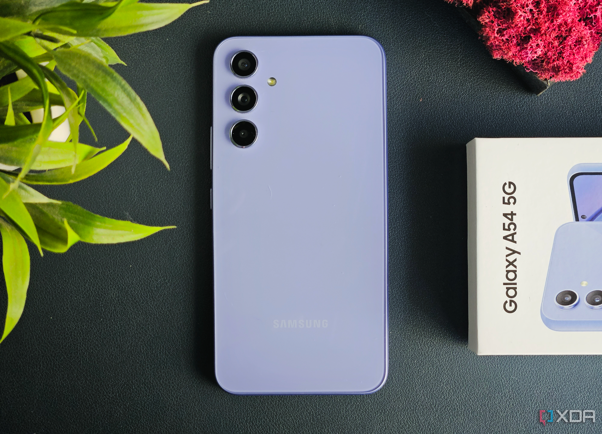 The Samsung Galaxy A54 in Awesome Violet color resting on a leather mat next to its retail box.