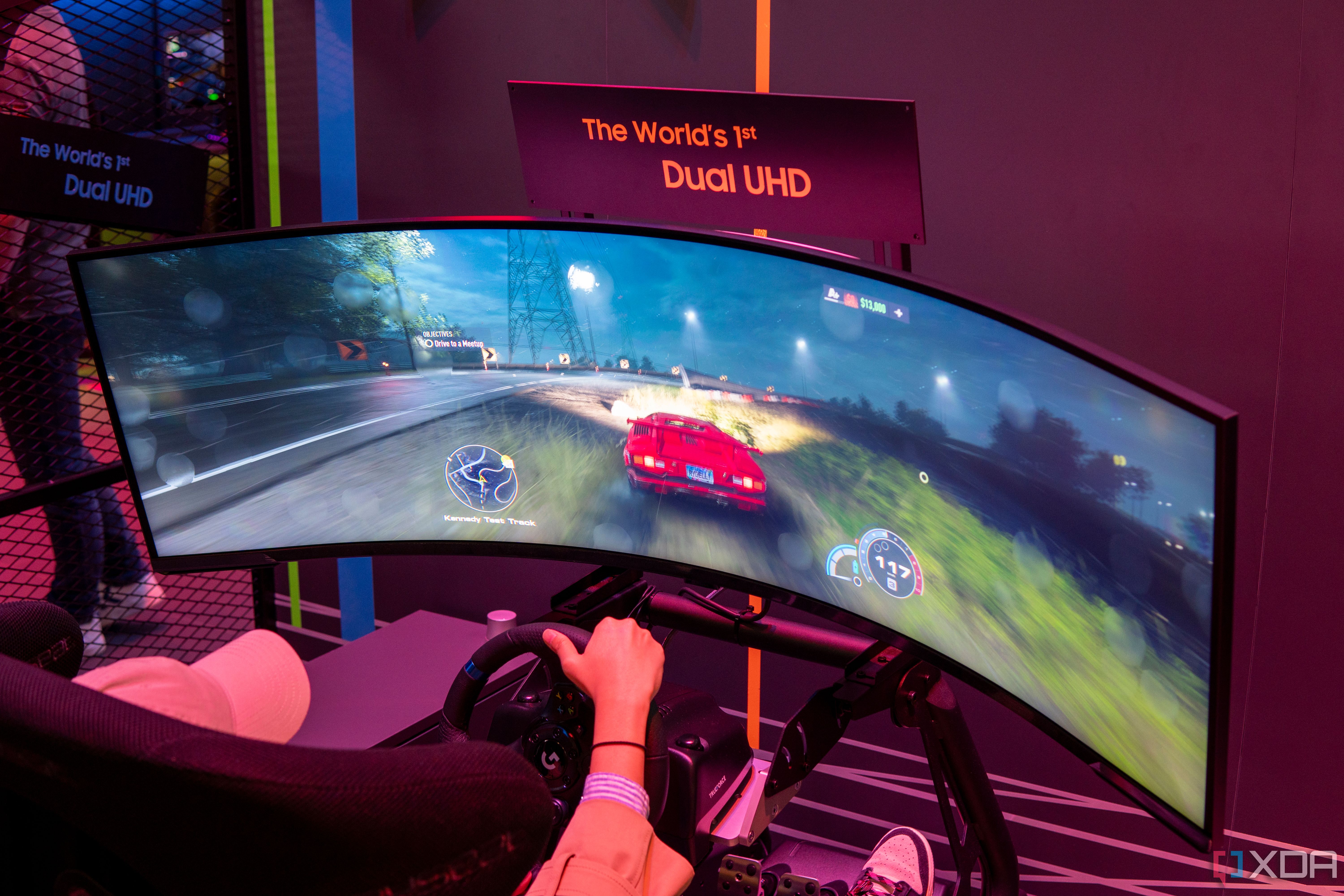 A Samsung Odyssey Neo G9 57-inch monitor displaying a racing game being played with a steering wheel.