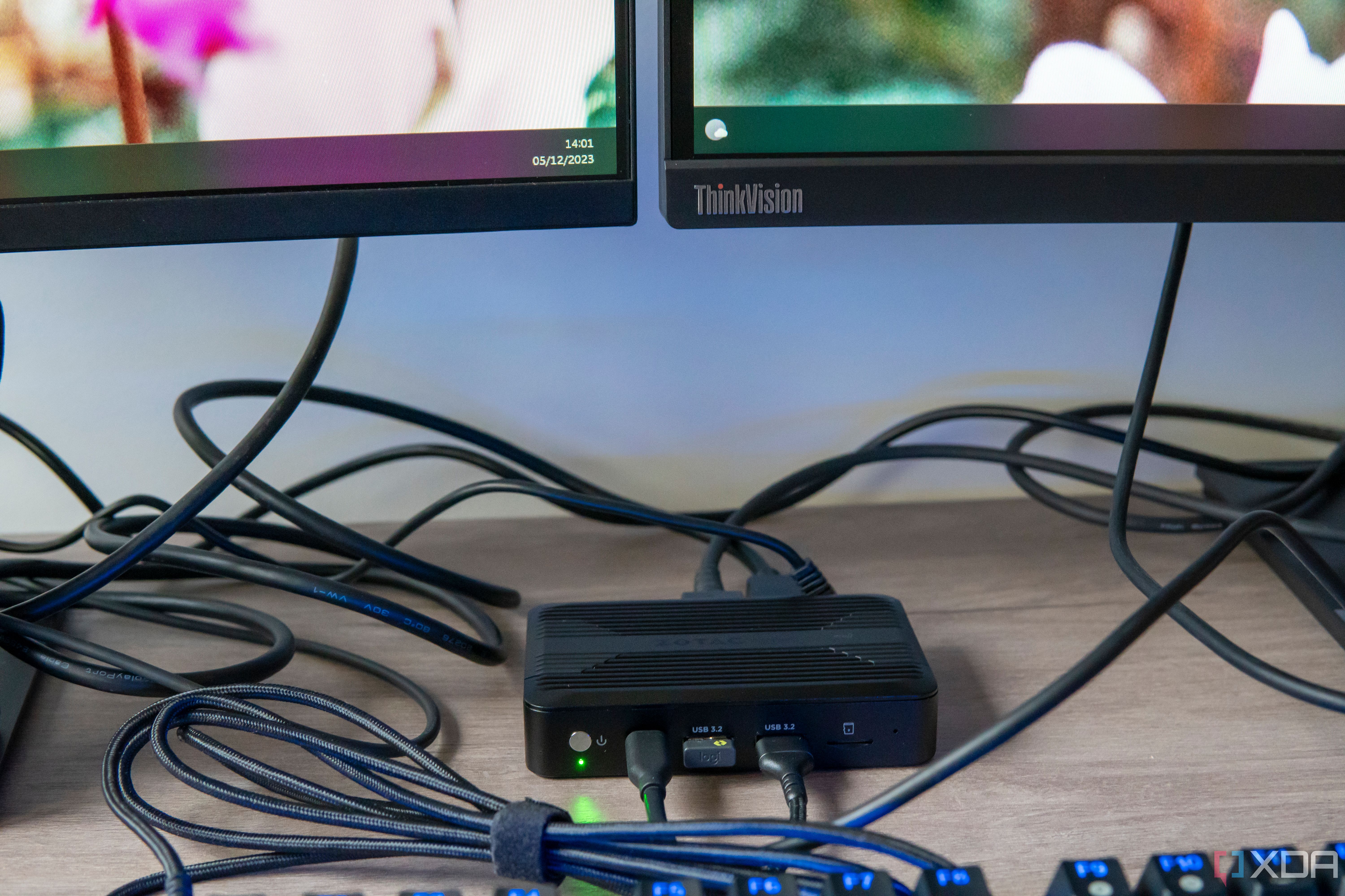 Angled front view of the Zotac Zbox Pico PI430AJ connected to multiple peripherals, including two monitors and a keyboard, which are visible in the frame