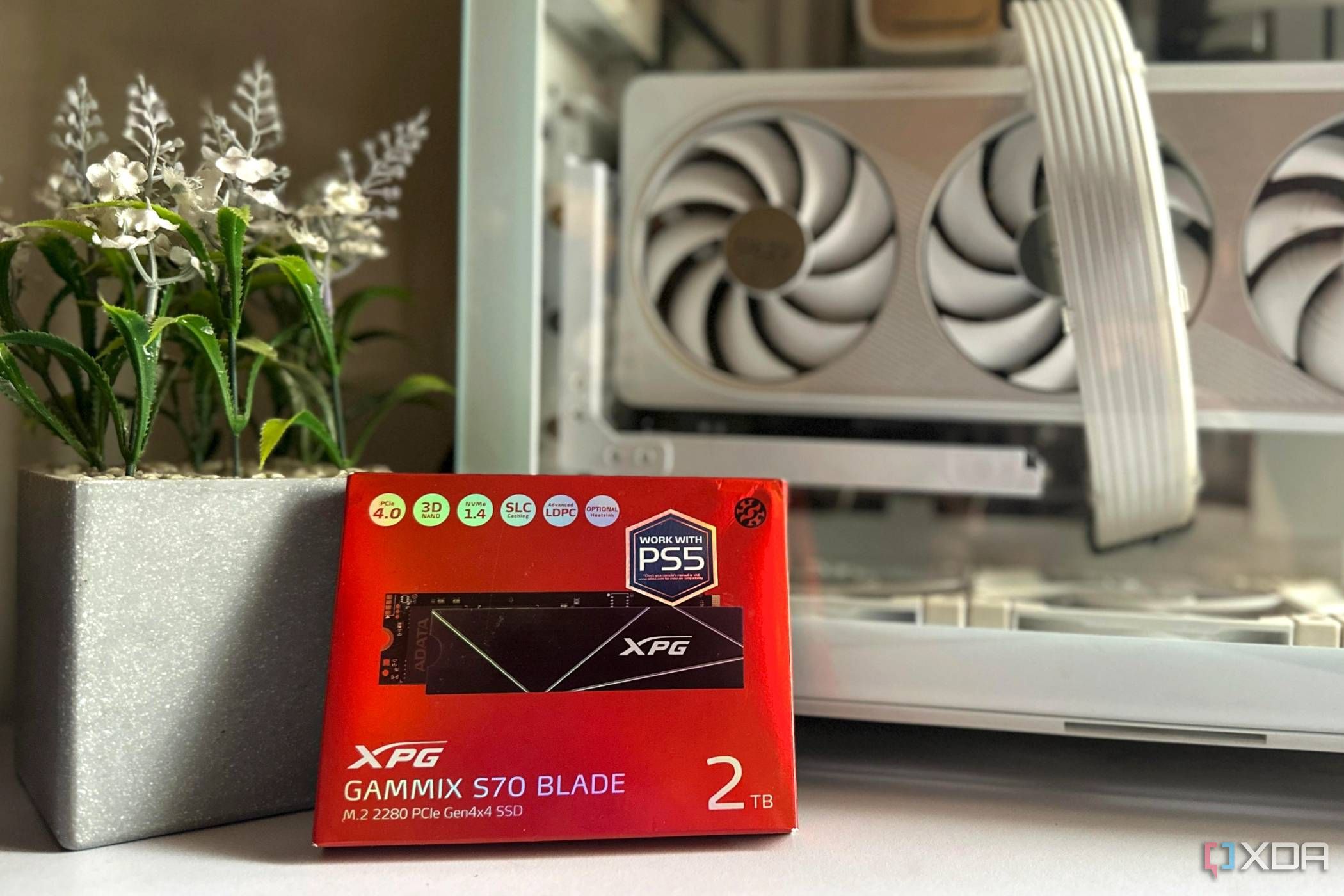 An image of a red box holding a 2TB SSD from XPG, resting against a faux flower pot and an unlit PC case